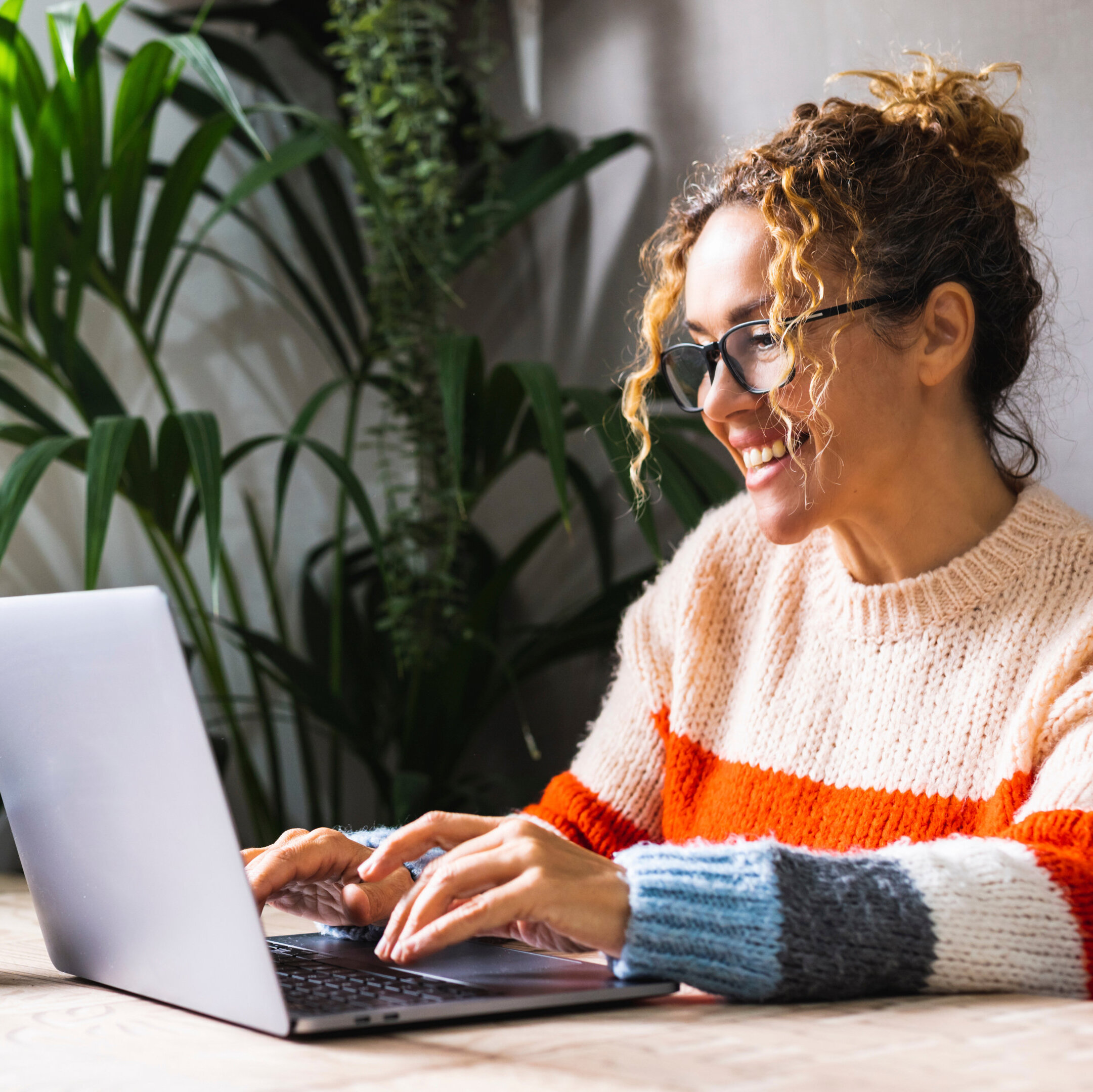 ächelnde Frau sitzt in einem Raum mit Pflanzen und arbeitet am Laptopiting on her laptop in a room with plants Lächelnde Frau sitzt in einem Raum mit Pflanzen und arbeitet am Laptop